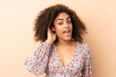 Genç Afrikalı Amerikalı bir kadın bej arka planda izole edilmiş bir şekilde elini kulağına koyarak bir şeyler dinliyor.