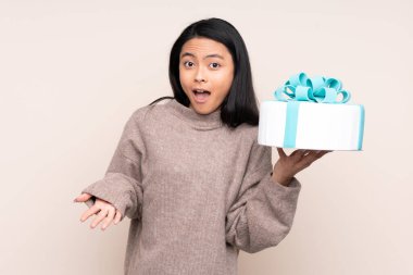 Genç Asyalı bir kız elinde büyük bir pasta tutuyor. Bej arka planda izole edilmiş ve yüzünde şok ifadesi var.