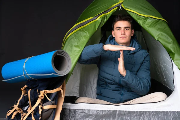 Teenager caucasian man inside a camping green tent isolated on black background making time out gesture