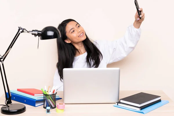 Student asian girl in a workplace with a laptop isolated on beige background making a selfie