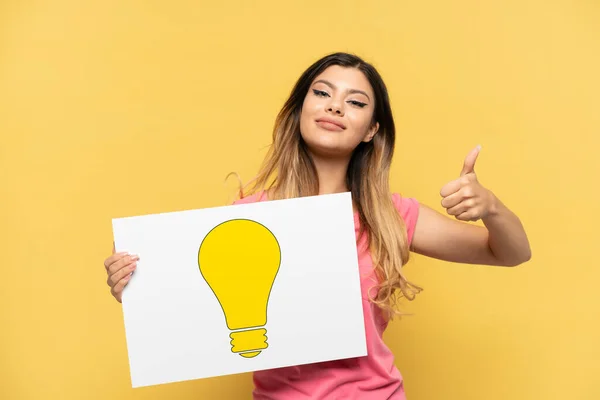 Young Russian Girl Isolated Yellow Background Holding Placard Bulb Icon — Stock Photo, Image