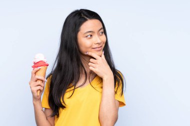 Kornet dondurması olan Asyalı genç bir kız mavi arka planda izole edilmiş bir şekilde bir fikir ve bir bakış açısı düşünüyor.