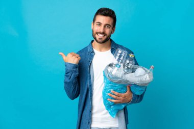 Mavi arka planda izole edilmiş plastik şişelerle dolu bir çanta taşıyan genç beyaz bir adam bir ürünü sunmak için kenarı işaret ediyor.