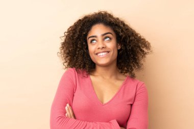 Genç Afrikalı Amerikalı kadın bej arka planda izole edilmiş gülümserken yukarı bakıyor