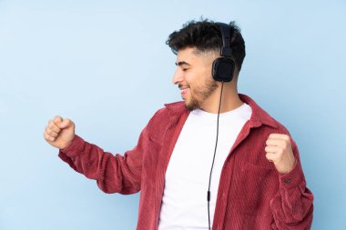 Arap yakışıklı adam mavi arka planda izole edilmiş müzik ve dans dinliyor.