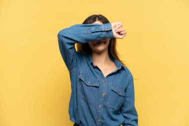 Sarı arka planda izole edilmiş genç bir Rus kız gözlerini elleriyle kapatıyor.