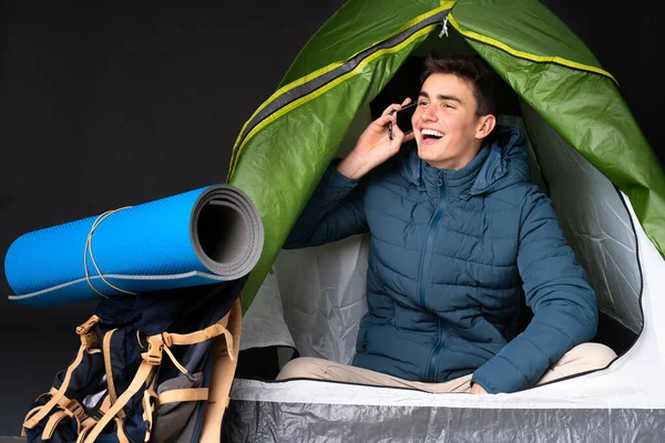 Teenager caucasian man inside a camping green tent isolated on black background keeping a conversation with the mobile phone
