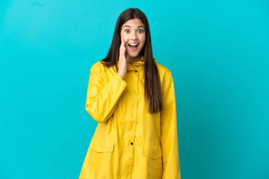 İzole edilmiş mavi arka planda yağmura dayanıklı bir ceket giyen Brezilyalı genç kız şaşırmış ve şok olmuş bir yüz ifadesi var.