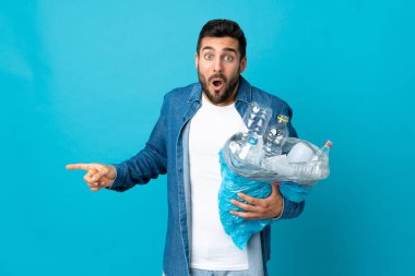 Mavi arka planda izole edilmiş plastik şişelerle dolu bir çanta taşıyan genç beyaz adam şaşırmış ve işaret ediyor.