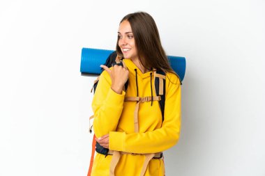 İzole edilmiş beyaz arka planı olan büyük bir sırt çantasıyla genç dağcı kadın bir ürünü sunmak için kenarı işaret ediyor.