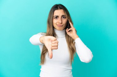 Mavi arka planda izole edilmiş cep telefonu kullanan genç beyaz kadın olumsuz ifade ile baş parmağını gösteriyor.