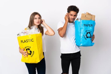 Beyaz arka planda izole edilmiş plastik ve kağıt dolu bir çanta taşıyan genç çift hayal kırıklığına uğramış ve kulaklarını kapatmış.