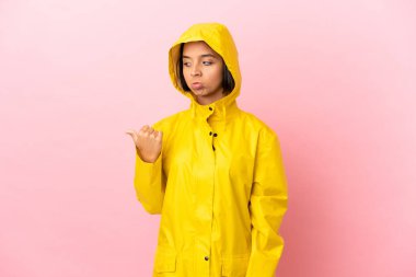 Yağmura dayanıklı bir ceket giyen genç Latin bir kadın izole edilmiş arka planda mutsuz ve bir tarafı işaret ediyor.