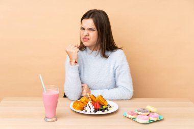 Bej arka planda izole edilmiş kızgın jestlerle waffle yiyen genç kız.