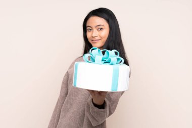 Genç Asyalı kız elinde büyük bir pasta tutuyor. Bej arka planda mutlu bir ifadeyle izole edilmiş.