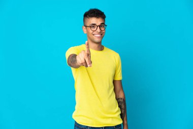 Mavi arka planda izole edilmiş genç Brezilyalı adam parmağını kaldırıyor ve gösteriyor.