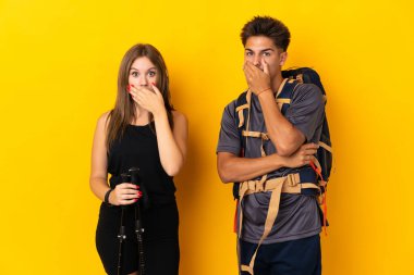 Sarı arka planda izole edilmiş büyük sırt çantalı genç dağcı çift uygunsuz bir şey söyledikleri için ağızlarını elleriyle kapatıyor.