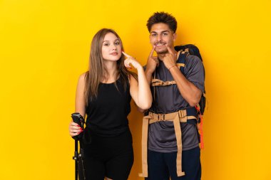 Sarı arka planda izole edilmiş büyük sırt çantalı genç dağcı çift mutlu ve hoş bir ifadeyle gülümsüyor.