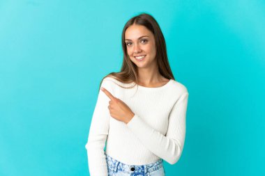 İzole edilmiş mavi arka planda genç bir kadın bir ürünü sunmak için kenarı işaret ediyor.