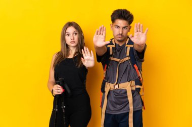Sarı arka planda izole edilmiş büyük sırt çantalı genç dağcı çift, bir görüşle hayal kırıklığına uğramak için dur hareketi yapıyorlar.