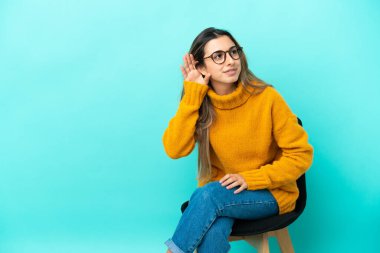 Mavi arka planda izole edilmiş bir sandalyede oturan beyaz bir kadın elini kulağına koyarak bir şeyler dinliyor.