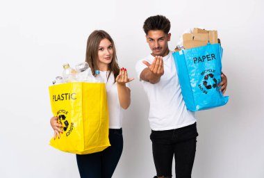 Beyaz arka planda izole edilmiş plastik ve kağıtlarla dolu bir çanta tutan genç çift el ele gelmeye davet ediyor. Gelmene sevindim.