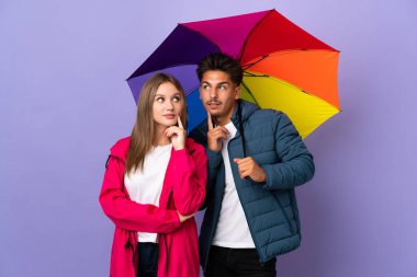 Mavi arka planda şemsiye tutan çift yukarı bakarken bir fikir düşünüyor.