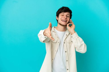 Mavi arka planda izole edilmiş cep telefonu kullanan genç bir Rus adam iyi bir anlaşma için el sıkışıyor.