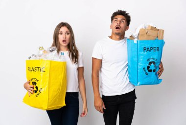 Beyaz arka planda izole edilmiş plastik ve kağıt dolu bir çanta taşıyan genç çift yukarı bakıyor ve şaşırmış bir ifadeyle