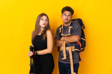 Sarı arka planda izole edilmiş büyük sırt çantalı genç dağcı çift omuzlarını kaldırırken şüpheyle hareket ediyorlar.