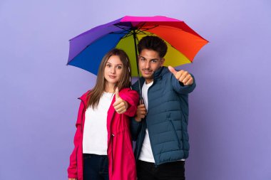 Mavi arka planda şemsiyesi olan çift iyi bir şey olduğu için baş parmak işareti yapıyor.