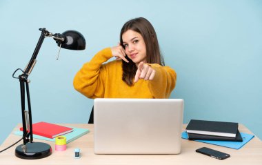 Öğrenci kız, mavi arka planda izole bir şekilde evinde çalışıyor. Telefon hareketleri yapıyor ve ön tarafı işaret ediyor.