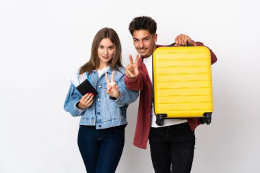 Mavi arka planda izole edilmiş bir bavul taşıyan gezgin çift gülümsüyor ve zafer işareti gösteriyor.