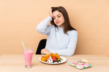 Bej arka planda izole edilmiş waffle yiyen genç kız hayal kırıklığı ifadesi sergiliyor ve anlamıyor.