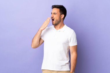 Mor arka planda soyutlanmış genç yakışıklı adam esniyor ve ağzını eliyle açık tutuyor.