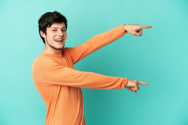 Genç Rus adam mavi arka planda izole edildi. Parmağını yana uzattı ve bir ürün sundu.