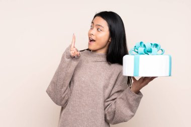 Genç Asyalı kız elinde bej arka planda izole edilmiş büyük bir pasta tutuyor. Çözümü bulmak için parmağını kaldırıyor.