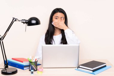 İşyerinde bej arka planda izole edilmiş dizüstü bilgisayarı olan Asyalı öğrenci ağzı elleriyle kapatıyor.