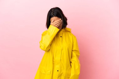 Yağmura dayanıklı bir ceket giyen genç Latin kadın izole bir arka plan ve yorgun ve hastalıklı bir yüz ifadesi var.