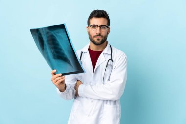 Radyografiyi elinde tutan profesyonel bir travmatolog mavi arka planda izole edilmiş, kollarını kavuşturuyor.