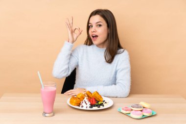 Bej arka planda izole edilmiş waffle yiyen genç kız şaşırdı ve tamam işareti gösteriyor.