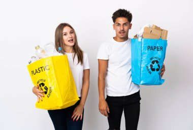 Plastik ve kağıt dolu bir çanta taşıyan genç bir çift beyaz arka planda şaşırtıcı bir yüz ifadesiyle izole edilmiş.