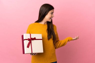 Genç Brezilyalı bir kız pembe arka planda bir hediye tutuyor. Sürpriz bir ifadeyle yan tarafa bakıyor.