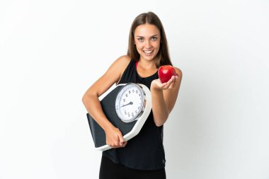 Soyutlanmış beyaz arka plan üzerinde genç bir kadın elinde tartı makinesi ve bir elma sunuyor.