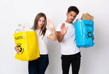 Beyaz arka planda izole edilmiş plastik ve kağıt dolu bir çanta taşıyan genç çift dur hareketi yapıyor ve hayal kırıklığına uğruyor.