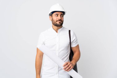 Miğferli ve elinde planları olan genç bir mimar beyaz arka planda izole edilmiş bir şekilde duruyor.