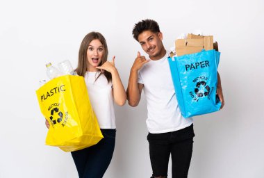 Beyaz arka planda izole edilmiş plastik ve kağıt dolu bir çanta taşıyan genç bir çift telefon jesti yapıyor. Bana geri dönüş işareti ver.
