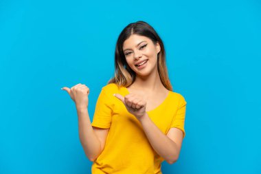 Mavi arka planda izole edilmiş genç bir Rus kız bir ürünü sunmak için kenarı işaret ediyor.