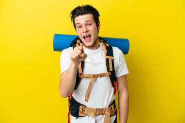 Sarı arka planda izole edilmiş büyük bir sırt çantası olan genç dağcı Rus adam şaşırmış ve ön tarafı işaret ediyor.