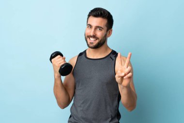 Sakallı genç sporcu halter kaldırıyor, gülümsüyor ve zafer işareti gösteriyor.